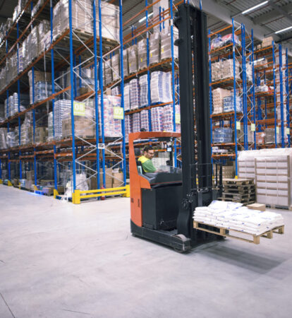 Worker operating forklift machine and relocating goods in large warehouse center.