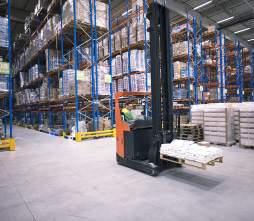 Worker operating forklift machine and relocating goods in large warehouse center.